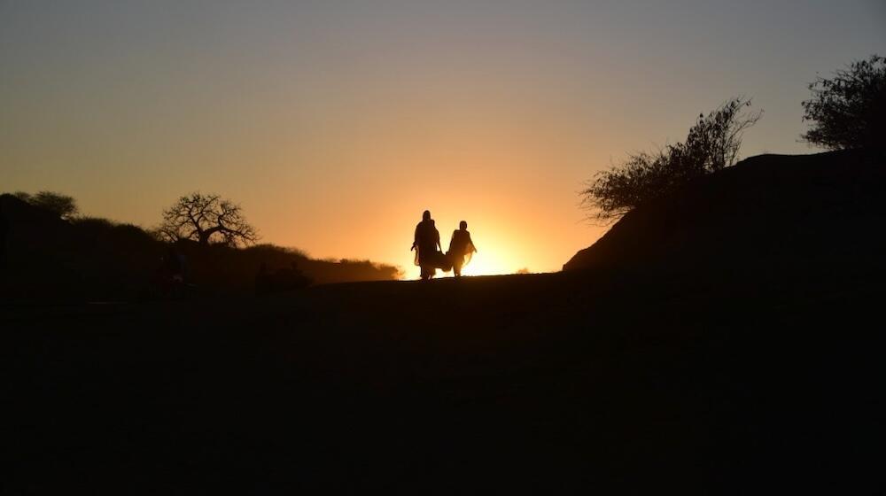 The image shows two figures walking along a path at sunset, silhouetted against the glowing horizon. The surrounding landscape is dark, with scattered bushes and trees framing the scene. The sunset bathes the sky in warm tones of orange and yellow, creating a serene and peaceful atmosphere. The figures appear to be walking together, symbolizing resilience, solidarity, or a journey toward hope amidst difficult circumstances.