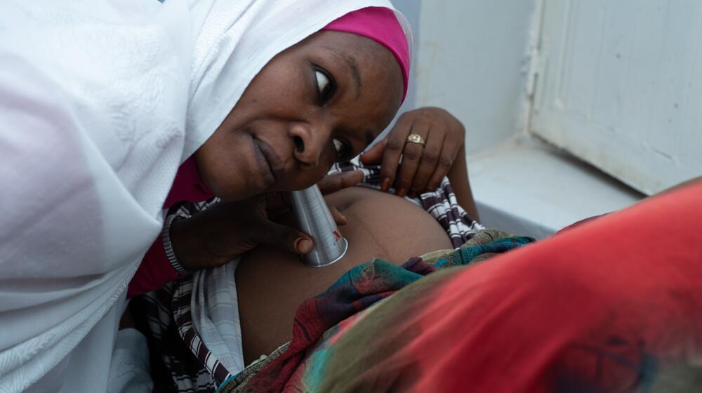 The image shows a midwife in Sudan attentively listening to the heartbeat of a pregnant woman using a fetal stethoscope. The midwife is wearing a white headscarf and focusing intently on providing care, highlighting the crucial role of health workers in ensuring safe pregnancies and deliveries, especially in conflict-affected regions.