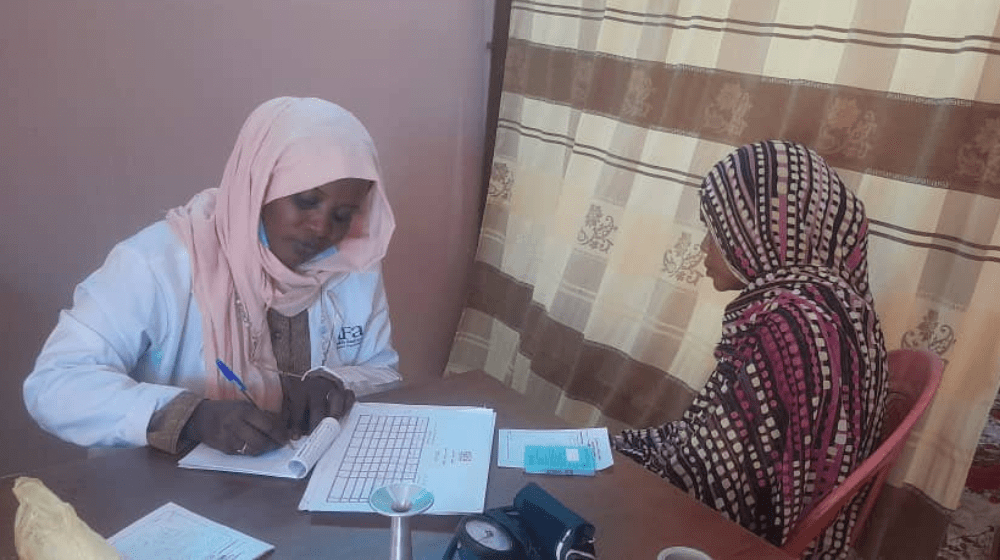 The image shows a healthcare worker wearing a white coat and a light pink headscarf sitting at a desk, writing in a medical logbook. Across from her sits a woman dressed in a patterned headscarf, facing the healthcare worker. They are in a modest clinic setting, separated from the rest of the room by a curtain. The scene reflects a consultation, where the healthcare worker is likely recording medical information while providing care to the woman, highlighting the importance of accessible healthcare services