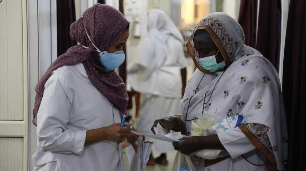 Two midwives at a hospital in Khartoum, Sudan, wearing protective masks, discuss documents and supplies. Despite the ongoing conflict, fuel shortages, and power cuts, they continue to provide critical maternal care, supported by UNFPA.