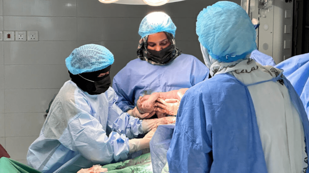  The image shows a group of healthcare professionals dressed in full surgical attire. They are in the process of delivering a newborn baby via Caesarean section in a medical facility. One of the professionals is holding the newborn, while others assist with the procedure. The environment appears to be sterile and well-equipped, with overhead surgical lights providing illumination. The healthcare workers’ focused expressions and careful handling of the newborn highlight the critical nature of their work.