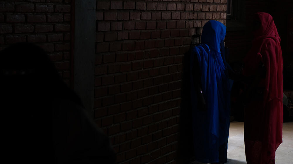 The image shows two women standing against a dimly lit brick wall, engaged in conversation. One woman is dressed in a blue robe, while the other wears a red robe. The shadows and low lighting create a sense of secrecy or caution, possibly reflecting the difficult and tense circumstances they are in. The setting suggests a private or hidden area, highlighting the challenges faced by women in conflict zones, where safety and privacy are often compromised.