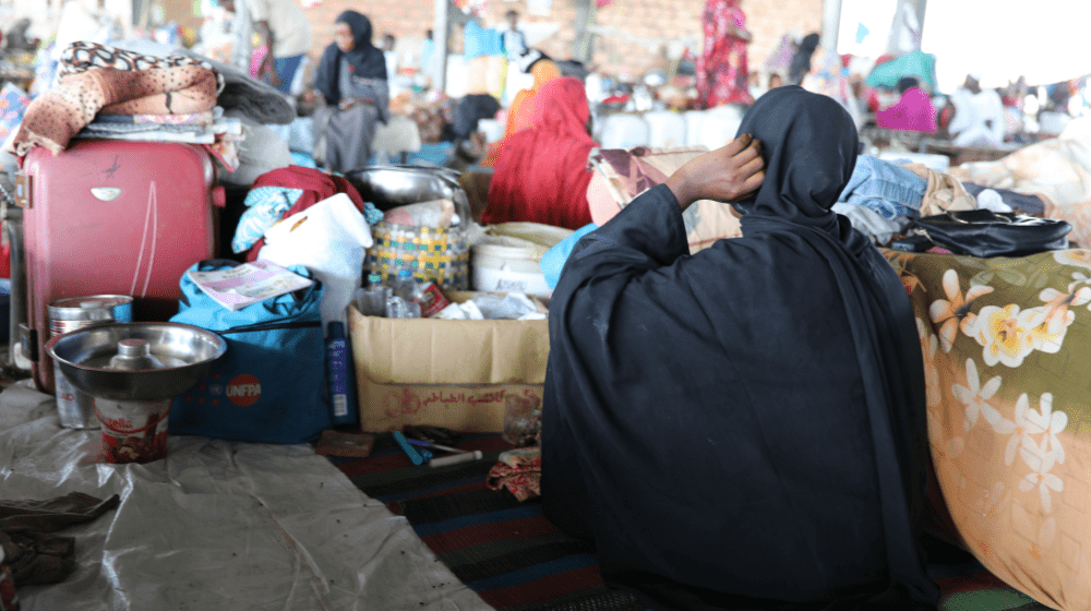  The image shows a woman sitting on the ground in a temporary shelter, surrounded by personal belongings, including blankets, bags, and household items. She is wearing a black headscarf and robe, with her back to the camera, adjusting her headscarf. Around her are scattered boxes, bags, and containers, including one with the UNFPA logo. The setting is crowded, with other people and belongings visible in the background, reflecting the difficult living conditions of displaced individuals in Sudan.